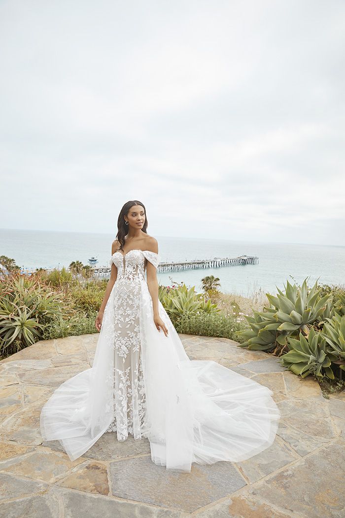 Ethereal and romantic, this off-the-shoulder lace gown features a sheer corset bodice, floral appliqués, and a detachabl