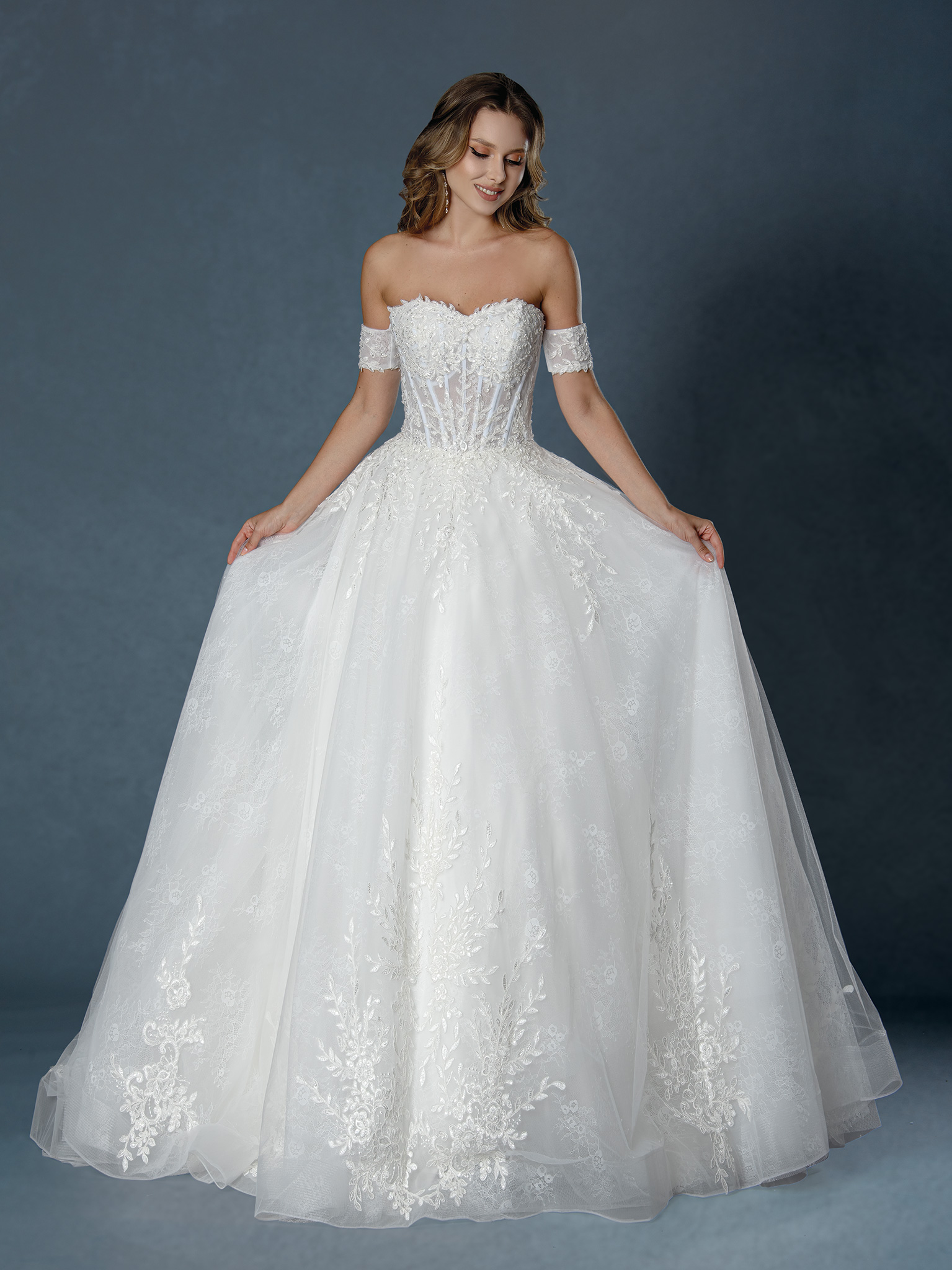 Whimsical and breathtaking, this off-the-shoulder ball gown features a sheer corset bodice, floral lace appliqués, and l
